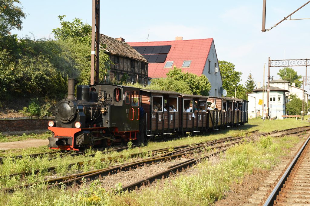 Kolejka wąskotorowa koloru czarno-czerwona z wagonami stoi na torach kolejowych.